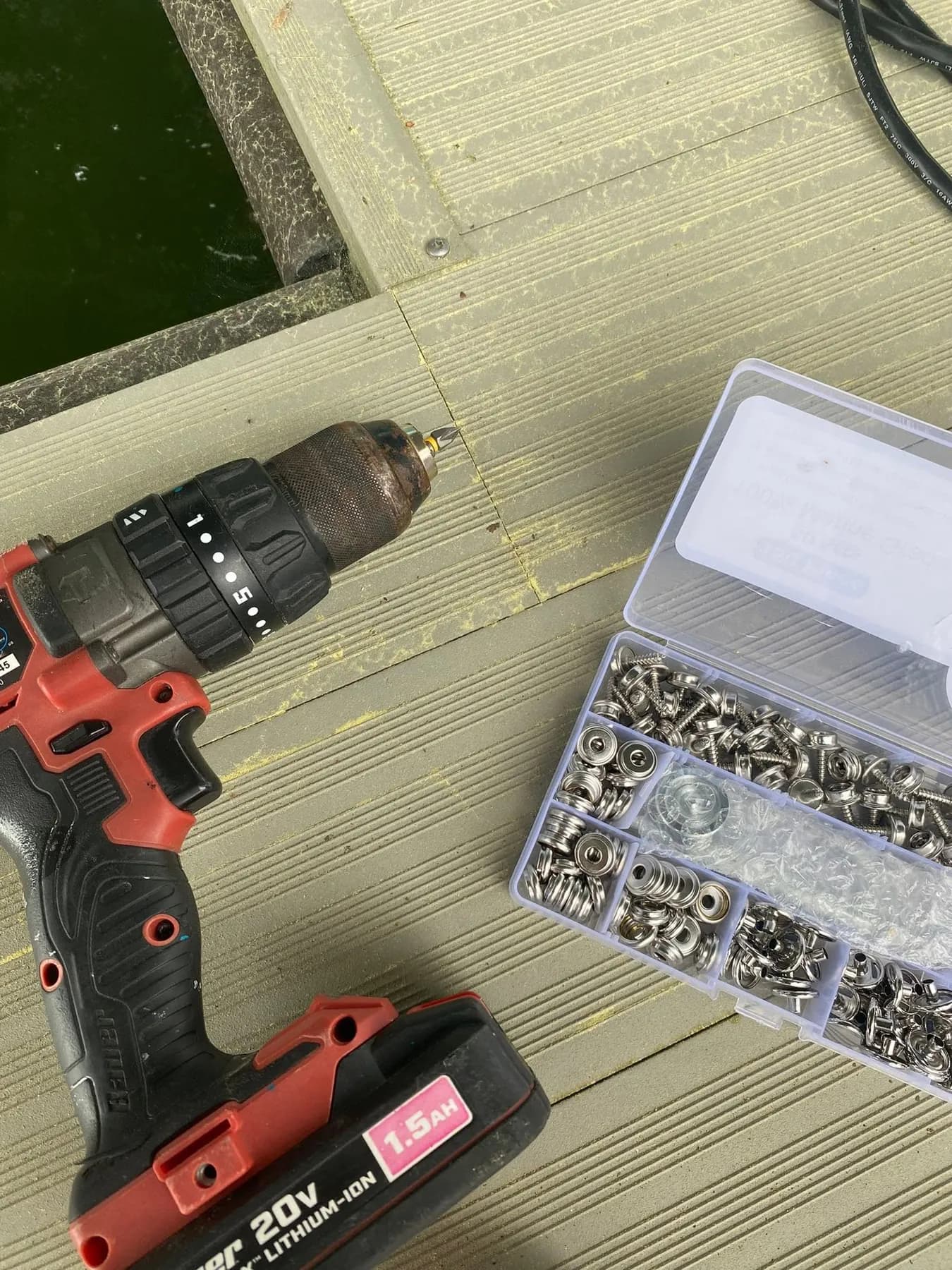 Cordless drill and a box of stainless snap fasteners on a dock above the water